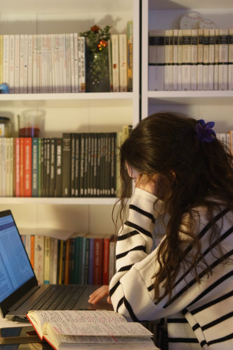 A woman in a striped sweater studies at home with a laptop and open notebook, surrounded by a bookshelf.